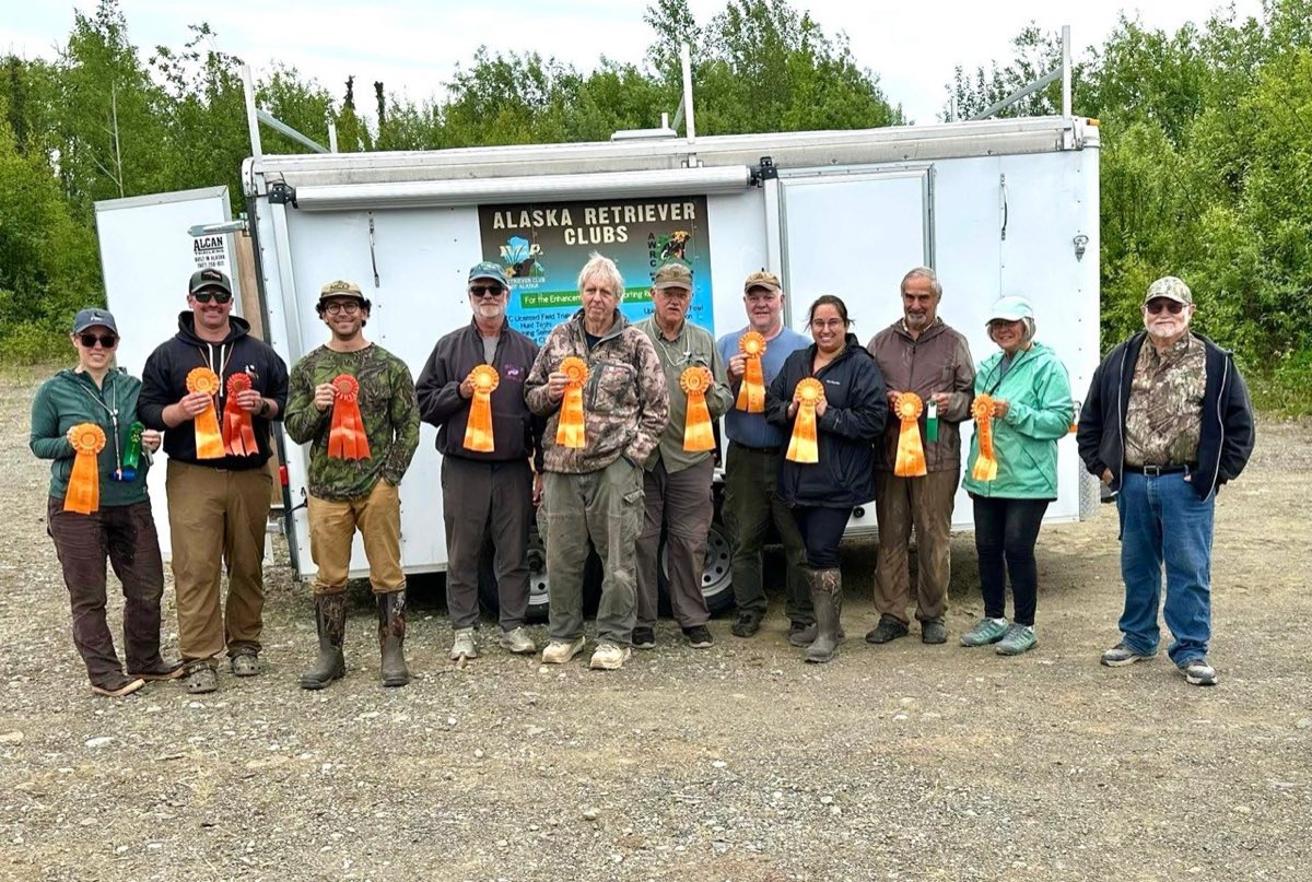 Twelve club members lined up with orange qualifying ribbons in front of the Alaska Retriever Clubs equipment trailer