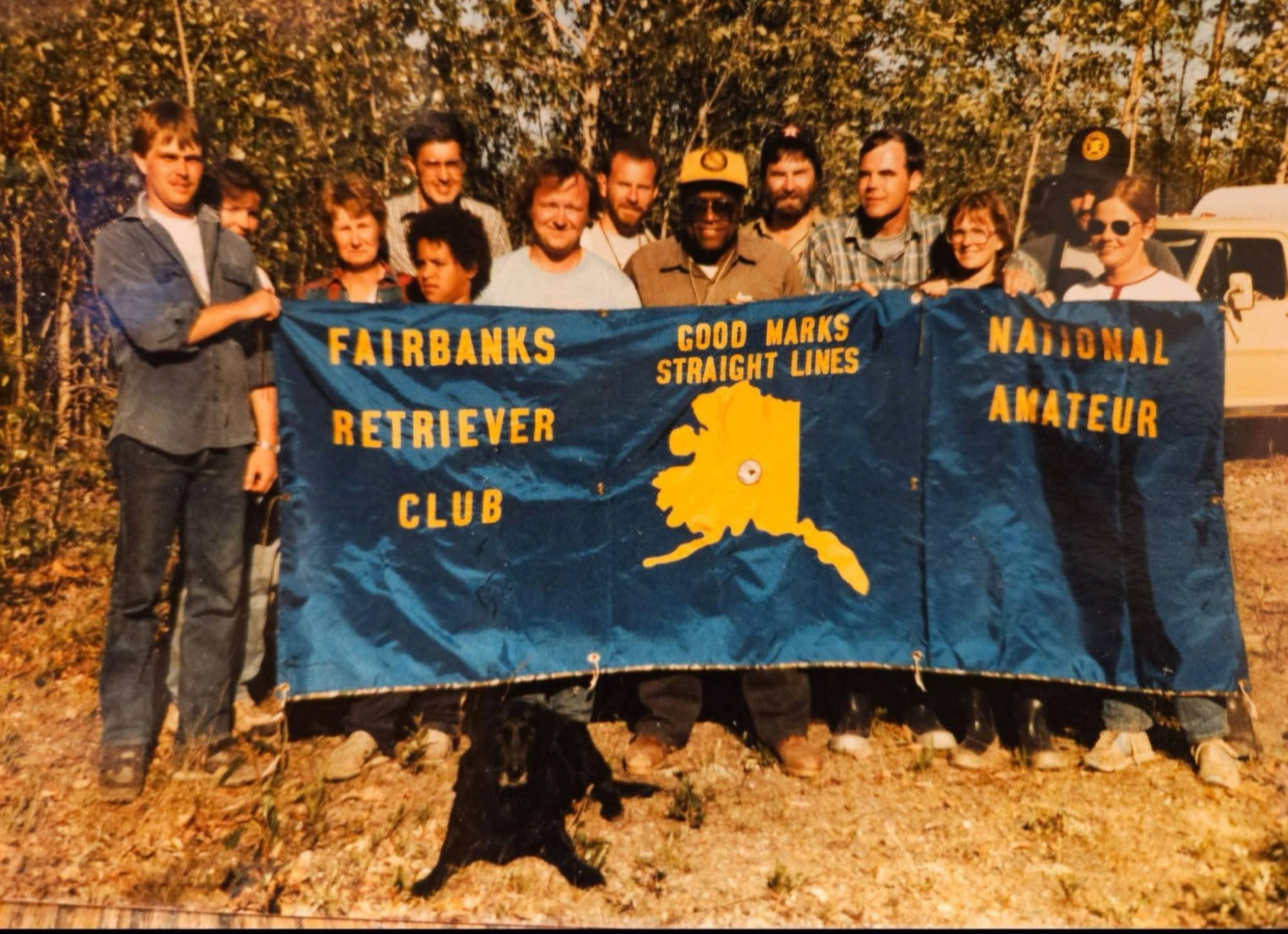 Around a dozen club members standing behind the club's blue holding blind, which reads Fairbanks Retriever Club, Good Marks Straight Lines and National Amateur around a gold outline of Alaska, with a black Labrador lying at their feet