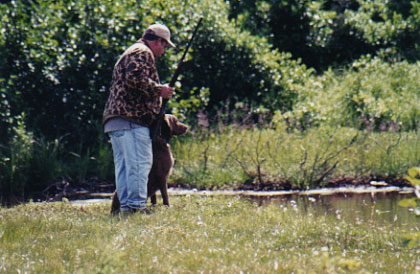 A handler standing at the water's edge with a Chesapeake Bay Retriever waiting beside him